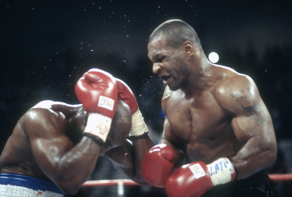 Evander Holyfield covers his ears with his gloves as Mike Tyson stands over him, baring his teeth.